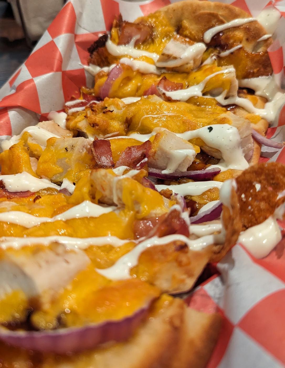A flatbread cut into slices in a red and white checkered paper basket, topped with chicken, bacon, red onion, melted cheddar and a white drizzle.