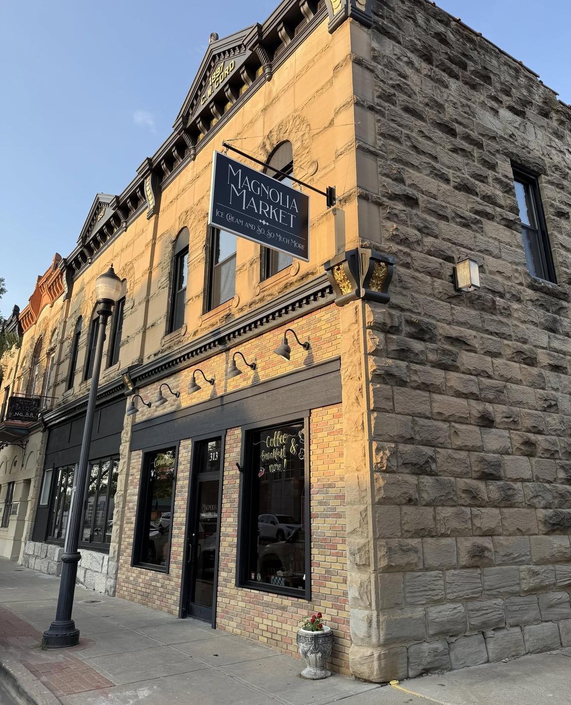 Magnolia Market on the square in Warrensburg: the corner stone and brick building with the hanging sign reading Magnolia Market, Ice Cream and So So Much More, the door numbered 313, gooseneck lights along the storefront and Coffee and Breakfast lettered on the window.
